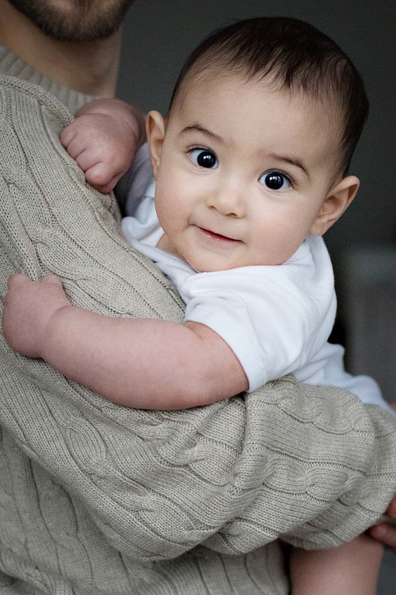 A baby with big brown eyes leaning over it's Dad's shoulder looking into camera