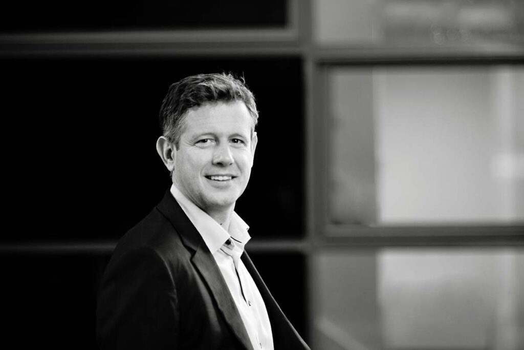 A corporate photo in black and white of a man in an open neck shirt with a jacket 